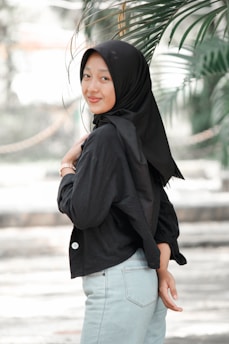 A young woman wearing a black hijab and a black jacket smiles while standing outdoors. She appears relaxed with palms and greenery in the background, suggesting a natural setting.