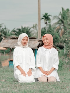 Two women are kneeling on grass in an outdoor setting with a rural backdrop. Both are wearing long white shirts; one wears a white headscarf while the other has a peach-colored headscarf. Palm trees and a wooden structure are visible in the background.