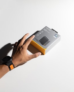 A hand wearing a black watch with an orange accent reaches for a product box labeled 'Voltme,' featuring images and descriptions of a charging device. The box has a gray and yellow color scheme and rests on a plain white surface, casting a shadow.