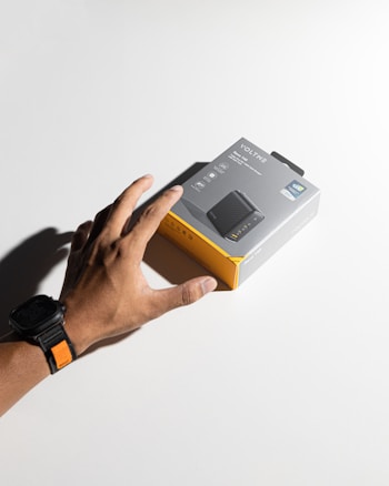 A hand wearing a black watch with an orange accent reaches for a product box labeled 'Voltme,' featuring images and descriptions of a charging device. The box has a gray and yellow color scheme and rests on a plain white surface, casting a shadow.