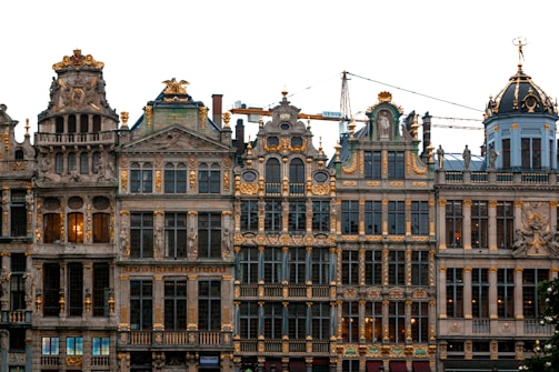 A row of ornate historic buildings with elaborate facades featuring intricate carvings and golden accents. The architecture showcases a blend of styles with prominent gables, arches, and decorative elements. A crane is visible in the background, hinting at ongoing construction or restoration.