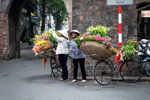 Two women wearing traditional conical hats are standing by bicycles filled with colorful flowers. They are positioned near a stone archway on a quiet street. One woman is adjusting flowers while the other holds a drink, both appear engaged in a casual moment.