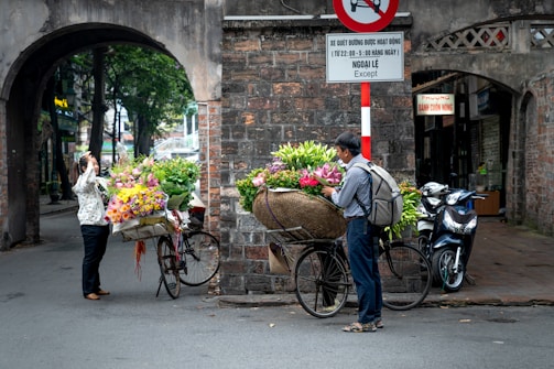 A street scene featuring two people beside bicycles loaded with vibrant flowers. One person is taking a photo of the flower arrangements while the other adjusts them. The backdrop includes an old brick archway and motorbikes parked nearby. A couple of signs are visible, adding to the urban setting.