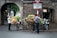 A street scene featuring two people beside bicycles loaded with vibrant flowers. One person is taking a photo of the flower arrangements while the other adjusts them. The backdrop includes an old brick archway and motorbikes parked nearby. A couple of signs are visible, adding to the urban setting.