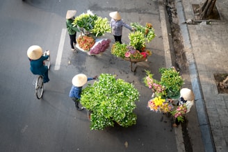 A group of people wearing traditional conical hats gather around vibrant bouquets and baskets filled with a variety of flowers. One person rides a bicycle, while others stand or walk on a paved street. The scene is captured from an overhead angle, showcasing the colorful floral arrangements and street setting.