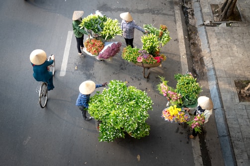 A group of people wearing traditional conical hats gather around vibrant bouquets and baskets filled with a variety of flowers. One person rides a bicycle, while others stand or walk on a paved street. The scene is captured from an overhead angle, showcasing the colorful floral arrangements and street setting.