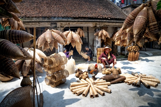 A marketplace scene displaying an array of woven bamboo products, including baskets and fish traps, arranged on the ground and hanging above. A person wearing a traditional hat sits among the items, with other individuals seen working in the background. The setting is a rustic courtyard or workshop, with weathered walls and a tiled roof, suggesting a traditional or rural environment.