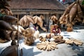 A marketplace scene displaying an array of woven bamboo products, including baskets and fish traps, arranged on the ground and hanging above. A person wearing a traditional hat sits among the items, with other individuals seen working in the background. The setting is a rustic courtyard or workshop, with weathered walls and a tiled roof, suggesting a traditional or rural environment.