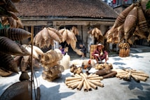 A marketplace scene displaying an array of woven bamboo products, including baskets and fish traps, arranged on the ground and hanging above. A person wearing a traditional hat sits among the items, with other individuals seen working in the background. The setting is a rustic courtyard or workshop, with weathered walls and a tiled roof, suggesting a traditional or rural environment.
