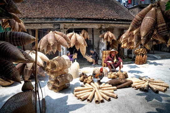 A marketplace scene displaying an array of woven bamboo products, including baskets and fish traps, arranged on the ground and hanging above. A person wearing a traditional hat sits among the items, with other individuals seen working in the background. The setting is a rustic courtyard or workshop, with weathered walls and a tiled roof, suggesting a traditional or rural environment.