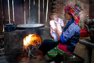 A woman dressed in colorful, traditional clothing holds a baby on her lap. Behind them, a small fire burns inside a rustic stove, with a black kettle placed on top. The setting appears to be a simple, wooden interior with brick walls.