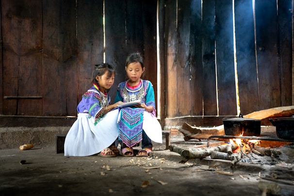 Two young girls are sitting together in a rustic interior, adorned in colorful traditional attire with intricate patterns. They are focused on a book, reading intently beside a simple fire. The setting is dimly lit, with light streaming through the wooden panels of the walls.
