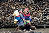 A woman and a child stand in front of a large stack of logs. The child carries a large plush toy, resembling a bear, wearing a red shirt and blue pants. Both the woman and child are wearing colorful, traditional clothing. They seem to be smiling and interacting warmly, suggesting a familial bond.