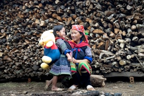A woman and a child stand in front of a large stack of logs. The child carries a large plush toy, resembling a bear, wearing a red shirt and blue pants. Both the woman and child are wearing colorful, traditional clothing. They seem to be smiling and interacting warmly, suggesting a familial bond.