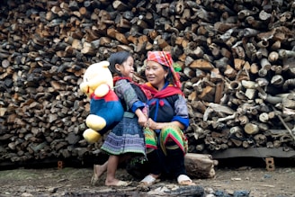 A woman and a child stand in front of a large stack of logs. The child carries a large plush toy, resembling a bear, wearing a red shirt and blue pants. Both the woman and child are wearing colorful, traditional clothing. They seem to be smiling and interacting warmly, suggesting a familial bond.