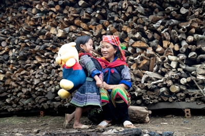 A woman and a child stand in front of a large stack of logs. The child carries a large plush toy, resembling a bear, wearing a red shirt and blue pants. Both the woman and child are wearing colorful, traditional clothing. They seem to be smiling and interacting warmly, suggesting a familial bond.