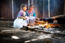 Two children in colorful, traditional clothing are sitting inside a rustic wooden structure. They are tending to a fire using sticks, and there is a black pot suspended over the flames. Smoke rises gently from the fire, and the room is dimly lit, with light filtering through gaps in the wooden walls.