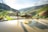 A vibrant landscape of terraced rice fields with lush greenery and hills in the background. Two people are present, one leading a water buffalo on a narrow path with water reflecting the bright sunlight. The scene is serene and picturesque, capturing the essence of rural life.