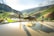 A vibrant landscape of terraced rice fields with lush greenery and hills in the background. Two people are present, one leading a water buffalo on a narrow path with water reflecting the bright sunlight. The scene is serene and picturesque, capturing the essence of rural life.