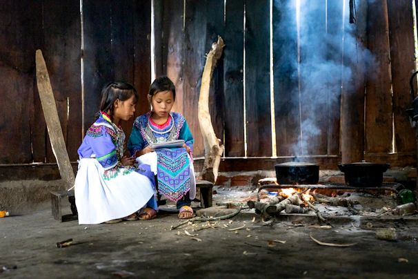 Two children dressed in colorful traditional clothing are seated together inside a rustic wooden structure. They are focused on something in their hands, while a small cooking fire with two pots emits a stream of smoke, creating a warm and cozy atmosphere.