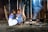 Two children dressed in colorful traditional clothing are seated together inside a rustic wooden structure. They are focused on something in their hands, while a small cooking fire with two pots emits a stream of smoke, creating a warm and cozy atmosphere.