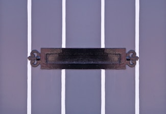 A close-up view of a decorative black mail slot on a gray paneled door. The door features vertical white stripes that contrast with the dark metal of the mail slot, which has intricate designs on its ends.