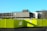 A multistory concrete parking structure under clear blue skies is seen behind a bright yellow construction barrier. The building has a stark, utilitarian appearance, and several leafless trees are visible nearby. The barrier displays directional signs and warning symbols.