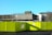 A multistory concrete parking structure under clear blue skies is seen behind a bright yellow construction barrier. The building has a stark, utilitarian appearance, and several leafless trees are visible nearby. The barrier displays directional signs and warning symbols.