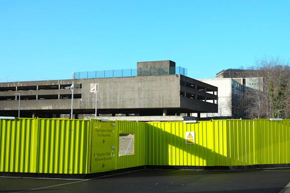 A multistory concrete parking structure under clear blue skies is seen behind a bright yellow construction barrier. The building has a stark, utilitarian appearance, and several leafless trees are visible nearby. The barrier displays directional signs and warning symbols.
