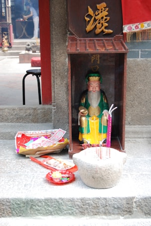 A colorful statue of a figure in traditional attire is placed inside a small wooden shrine with gold lettering. In front of the statue, there is a stone container holding several incense sticks. On the ground nearby, there are various offerings including paper money, fruit, and packaged food items. Smoke from the incense is faintly visible, contributing to a serene atmosphere.