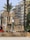 Large illuminated sign reading 'Gran Fira de Valencia' stands prominently in an urban setting surrounded by palm trees and multistory buildings. The sign features vintage-style light bulbs and is situated near a busy street with parked cars and a red bus in the background.