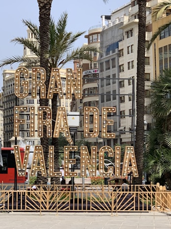 Large illuminated sign reading 'Gran Fira de Valencia' stands prominently in an urban setting surrounded by palm trees and multistory buildings. The sign features vintage-style light bulbs and is situated near a busy street with parked cars and a red bus in the background.