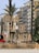 Large illuminated sign reading 'Gran Fira de Valencia' stands prominently in an urban setting surrounded by palm trees and multistory buildings. The sign features vintage-style light bulbs and is situated near a busy street with parked cars and a red bus in the background.