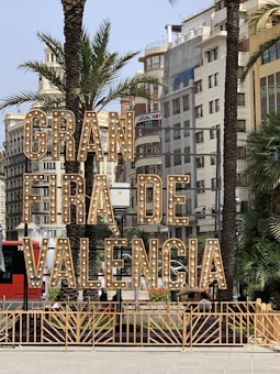 Large illuminated sign reading 'Gran Fira de Valencia' stands prominently in an urban setting surrounded by palm trees and multistory buildings. The sign features vintage-style light bulbs and is situated near a busy street with parked cars and a red bus in the background.
