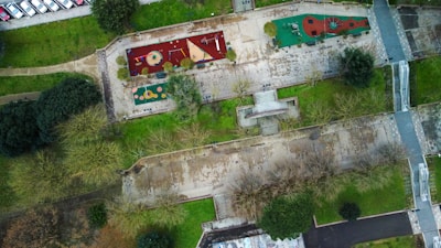 Aerial view of a park featuring two playground areas with various equipment like slides and climbing structures. The playgrounds have colorful surfaces, surrounded by green grass and trees. Pathways and a staircase connect different parts of the park, with a line of parked cars visible on the left side.