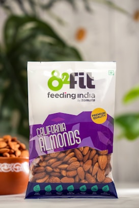 A clear plastic bag containing California almonds is prominently displayed against a blurred background with greenery. The packaging includes branding with the logo '82fit feeding india by zomato' and a purple design with health benefits and 'premium quality' label. A bowl filled with almonds is partially visible on the left.