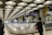 A modern airport baggage claim area with a highly structured ceiling of circular light panels. The conveyor belt is empty, and a few people are visible in the background, holding luggage. Tall pillars and signs with yellow accents are present, creating an industrial yet clean appearance.
