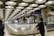 A modern airport baggage claim area with a highly structured ceiling of circular light panels. The conveyor belt is empty, and a few people are visible in the background, holding luggage. Tall pillars and signs with yellow accents are present, creating an industrial yet clean appearance.