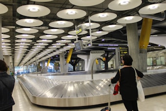 A modern airport baggage claim area with a highly structured ceiling of circular light panels. The conveyor belt is empty, and a few people are visible in the background, holding luggage. Tall pillars and signs with yellow accents are present, creating an industrial yet clean appearance.