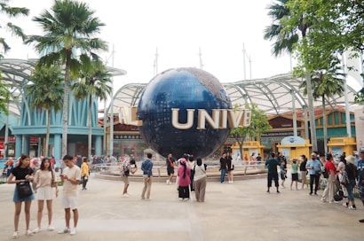 A large globe structure with the letters 'UNIVE' visible, surrounded by a crowd of people taking photos and walking around. The area is lush with palm trees and modern architectural elements, possibly in an amusement or theme park setting.