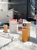 A parking payment kiosk and signage are partially surrounded by snow in an urban environment. The building backdrop is composed of grey concrete with modern architectural lines. Various signs display information in Japanese, with one indicating parking rates. The scene is illuminated by bright sunlight, casting distinct shadows on the snow-covered ground.