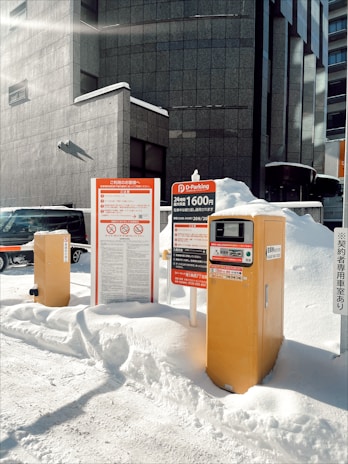 A parking payment kiosk and signage are partially surrounded by snow in an urban environment. The building backdrop is composed of grey concrete with modern architectural lines. Various signs display information in Japanese, with one indicating parking rates. The scene is illuminated by bright sunlight, casting distinct shadows on the snow-covered ground.