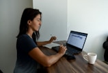 A woman sits at a wooden table working on a laptop. She is holding a pen and reviewing something on the screen. A notebook is positioned next to the laptop. A white coffee mug is placed on the table. The setting suggests a quiet indoor workspace with natural light illuminating the scene.