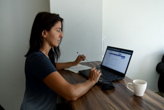 A woman sits at a wooden table working on a laptop. She is holding a pen and reviewing something on the screen. A notebook is positioned next to the laptop. A white coffee mug is placed on the table. The setting suggests a quiet indoor workspace with natural light illuminating the scene.