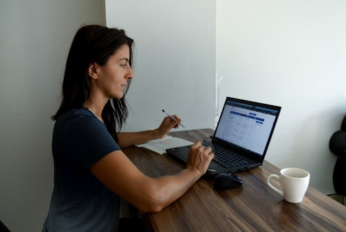 A woman sits at a wooden table working on a laptop. She is holding a pen and reviewing something on the screen. A notebook is positioned next to the laptop. A white coffee mug is placed on the table. The setting suggests a quiet indoor workspace with natural light illuminating the scene.
