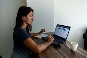 A woman sits at a wooden table working on a laptop. She is holding a pen and reviewing something on the screen. A notebook is positioned next to the laptop. A white coffee mug is placed on the table. The setting suggests a quiet indoor workspace with natural light illuminating the scene.