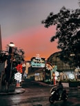 An illuminated arch with festive decorations and the message 'Have a New Destination King Power Celebrations 2023' is situated on a street. The sky transitions to a warm, glowing hue of pink and orange at sunset, while city buildings surround the scenery. A person on a motorcycle moves through the foreground, and decorative lights include stars and snowflakes.
