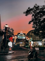An illuminated arch with festive decorations and the message 'Have a New Destination King Power Celebrations 2023' is situated on a street. The sky transitions to a warm, glowing hue of pink and orange at sunset, while city buildings surround the scenery. A person on a motorcycle moves through the foreground, and decorative lights include stars and snowflakes.