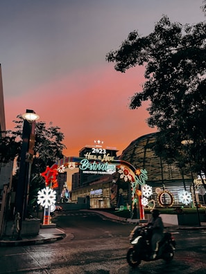 An illuminated arch with festive decorations and the message 'Have a New Destination King Power Celebrations 2023' is situated on a street. The sky transitions to a warm, glowing hue of pink and orange at sunset, while city buildings surround the scenery. A person on a motorcycle moves through the foreground, and decorative lights include stars and snowflakes.