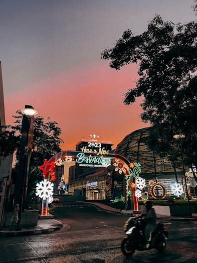 An illuminated arch with festive decorations and the message 'Have a New Destination King Power Celebrations 2023' is situated on a street. The sky transitions to a warm, glowing hue of pink and orange at sunset, while city buildings surround the scenery. A person on a motorcycle moves through the foreground, and decorative lights include stars and snowflakes.